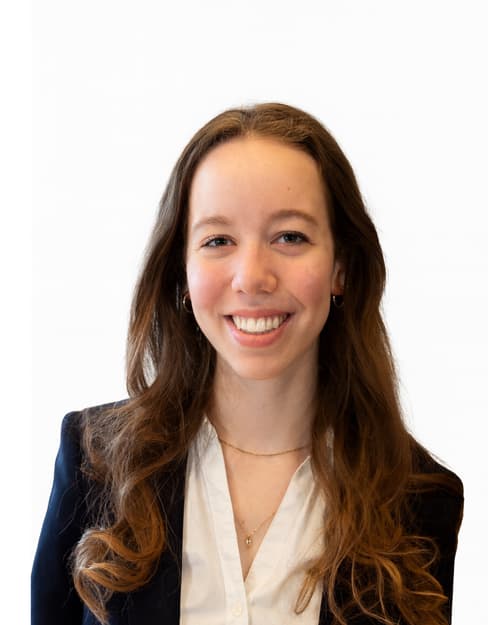 Young woman with long brown curly hair smiling, wearing a black blazer, white blouse, gold necklaces, and small hoop earrings, standing against a plain white background.