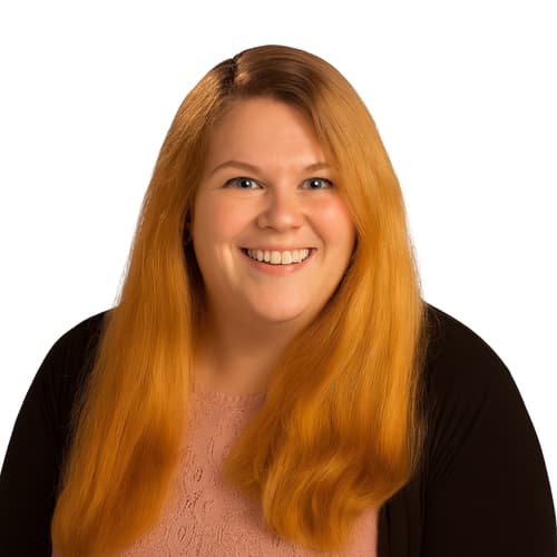 Portrait of a woman with long red hair, blue eyes, and a friendly smile, wearing a pink lace top and a black cardigan, against a white background.