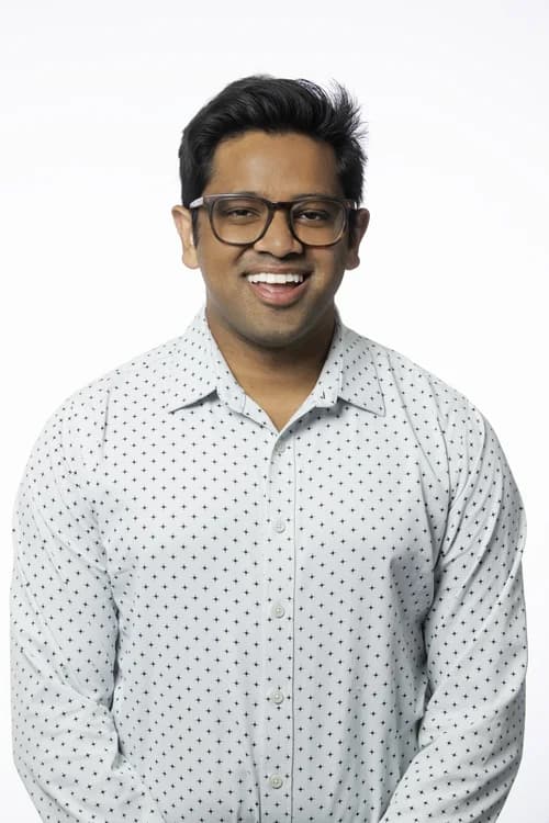 A man wearing glasses and a white shirt with small black dots, smiling against a plain white background.