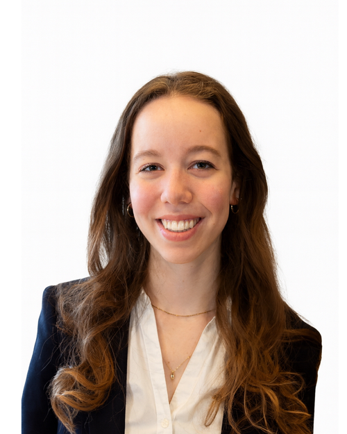 Young woman with long brown curly hair smiling, wearing a black blazer, white blouse, gold necklaces, and small hoop earrings, standing against a plain white background.