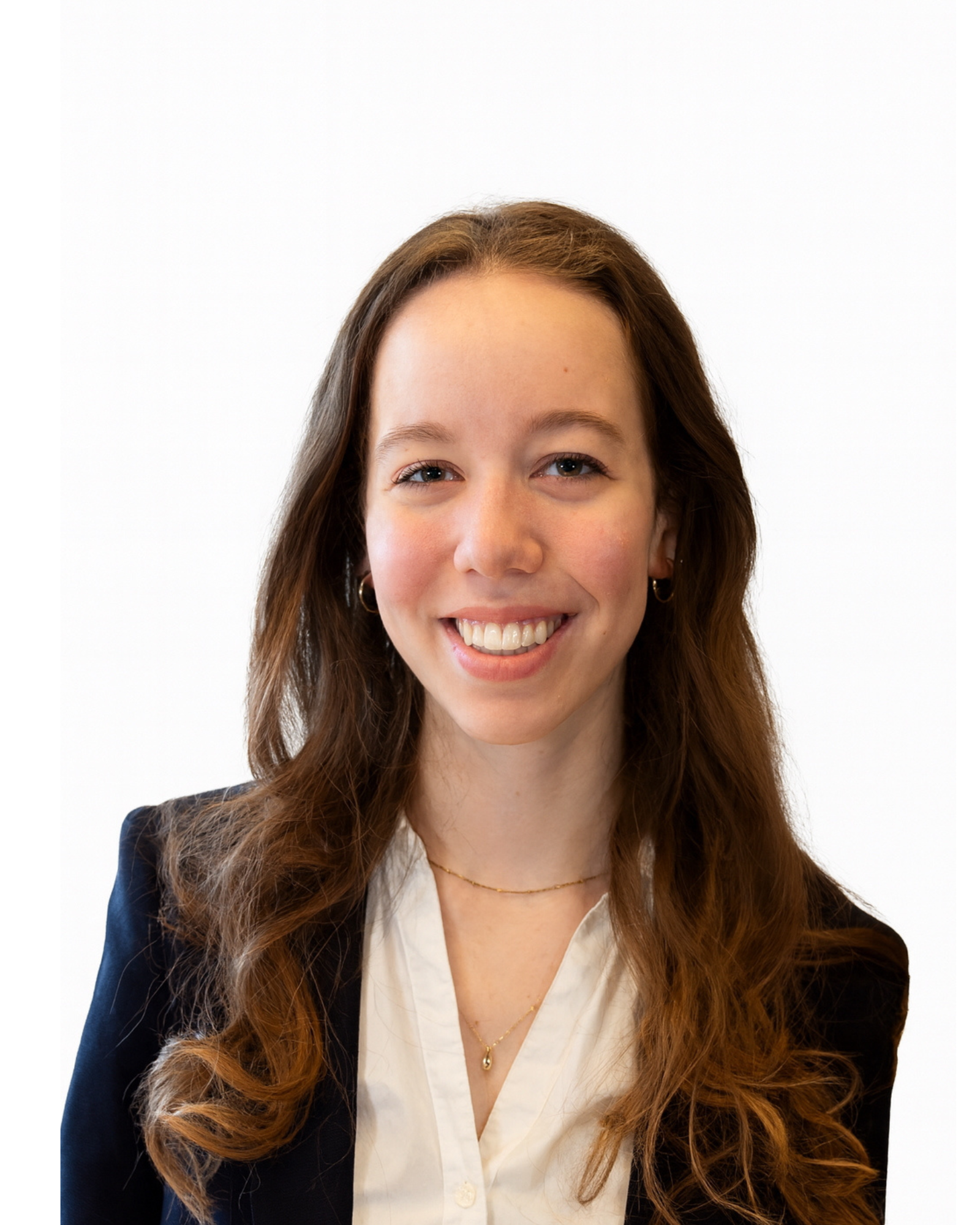 Young woman with long brown curly hair smiling, wearing a black blazer, white blouse, gold necklaces, and small hoop earrings, standing against a plain white background.