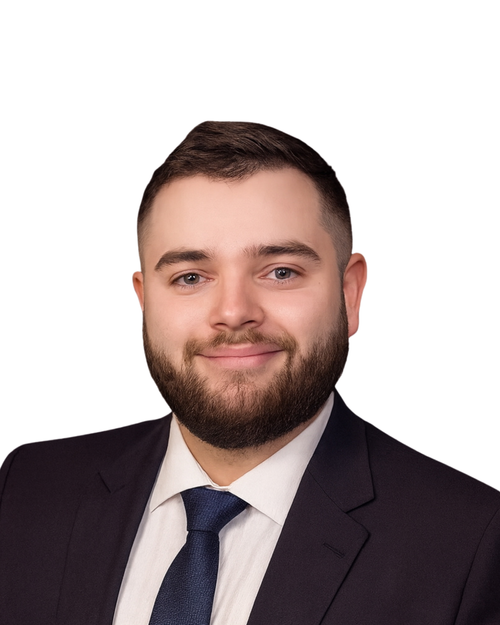 A professional headshot of a young man with short brown hair, a well-groomed beard, wearing a dark suit, white shirt, and navy tie, smiling against a plain white background.