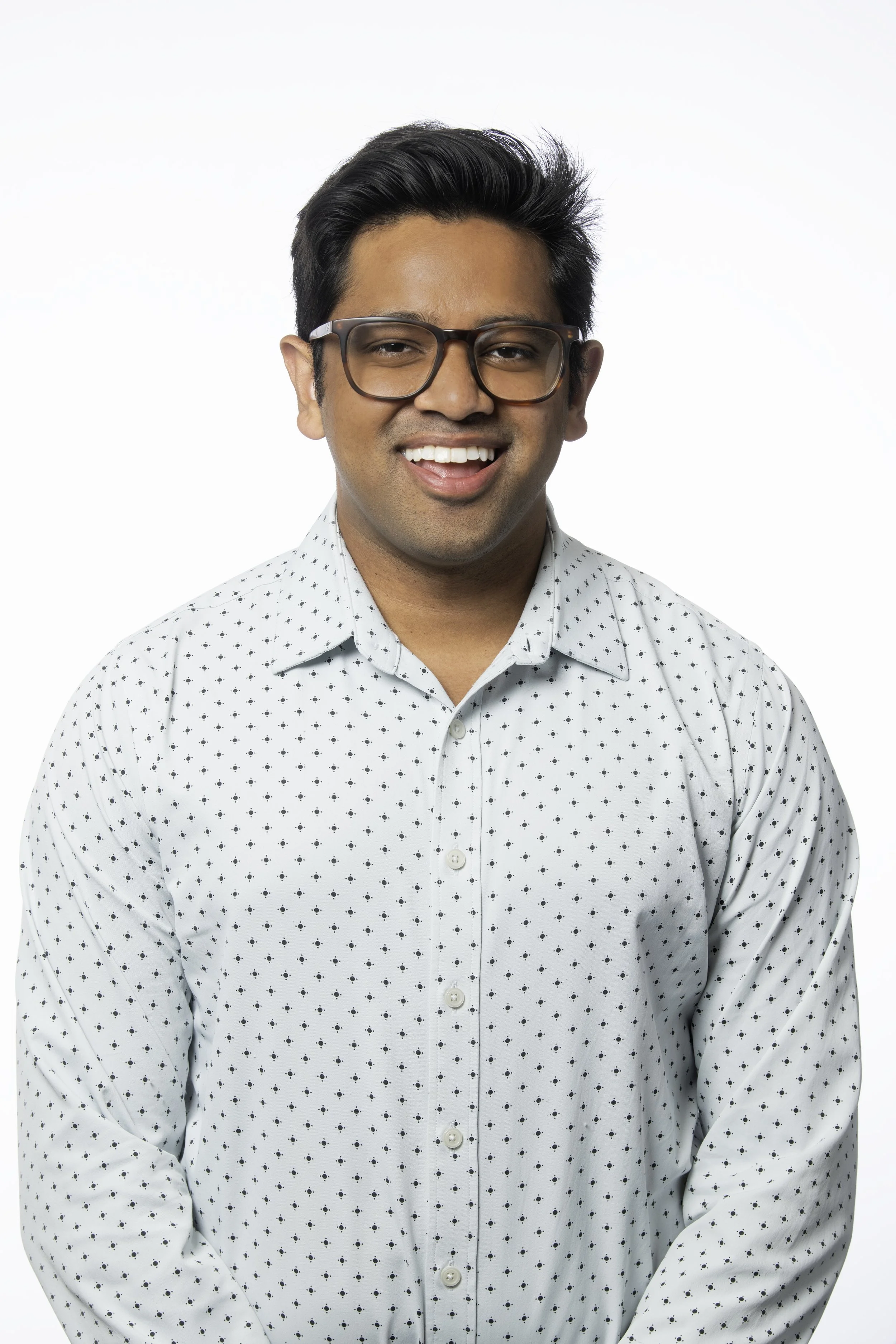 A man wearing glasses and a white shirt with small black dots, smiling against a plain white background.